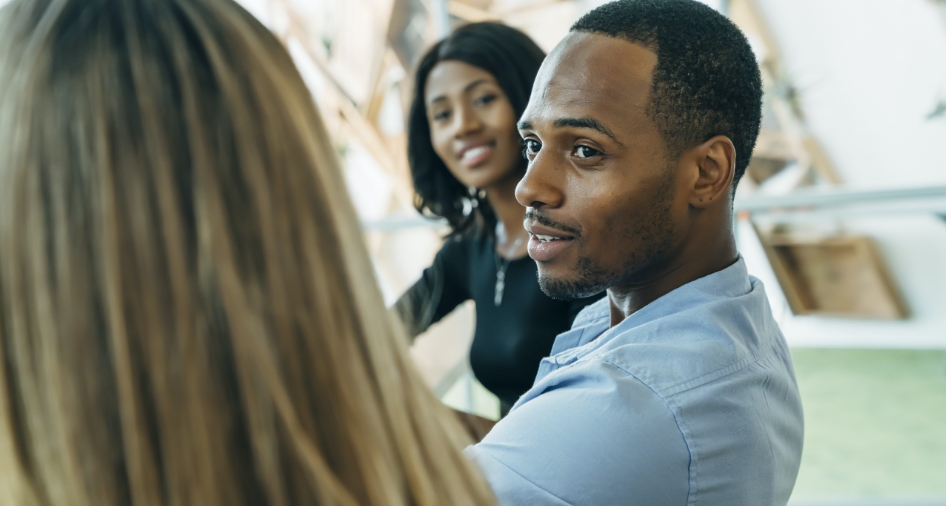 This image shows 3 people. Two women and a man in the middle of the three. They are all sitting down having a discussion. The man is  speaking to the woman on his left with the woman on his right listening in to the conversation