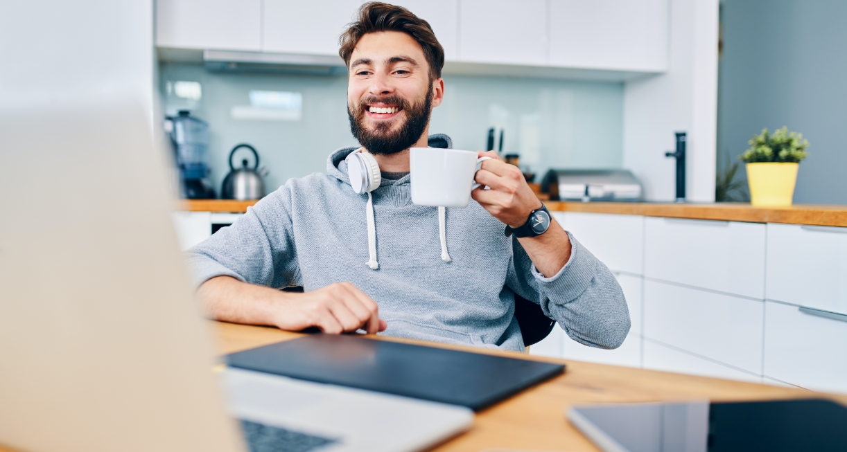 This images showcases a middle aged man sitting down in his kitchen and having a virtual meeting. He is smiling and holds a mug in his hands