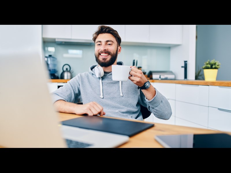 This images showcases a middle aged man sitting down in his kitchen and having a virtual meeting. He is smiling and holds a mug in his hands